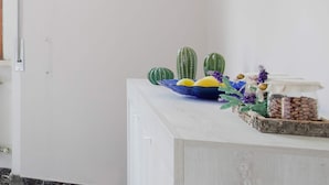 Un meuble blanc surmonté d’un saladier bleu contenant des citrons et une pastèque verte, ainsi que deux cactus et une composition florale décorative.