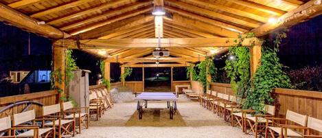 A covered outdoor seating area with wooden chairs and tables, a gravel floor, and wooden beams on the ceiling.