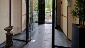 Um corredor com porta de vidro que dá para um pátio. Há uma pequena mesa com um vaso e uma estátua à esquerda. O chão é ladrilhado e há placas nas paredes.