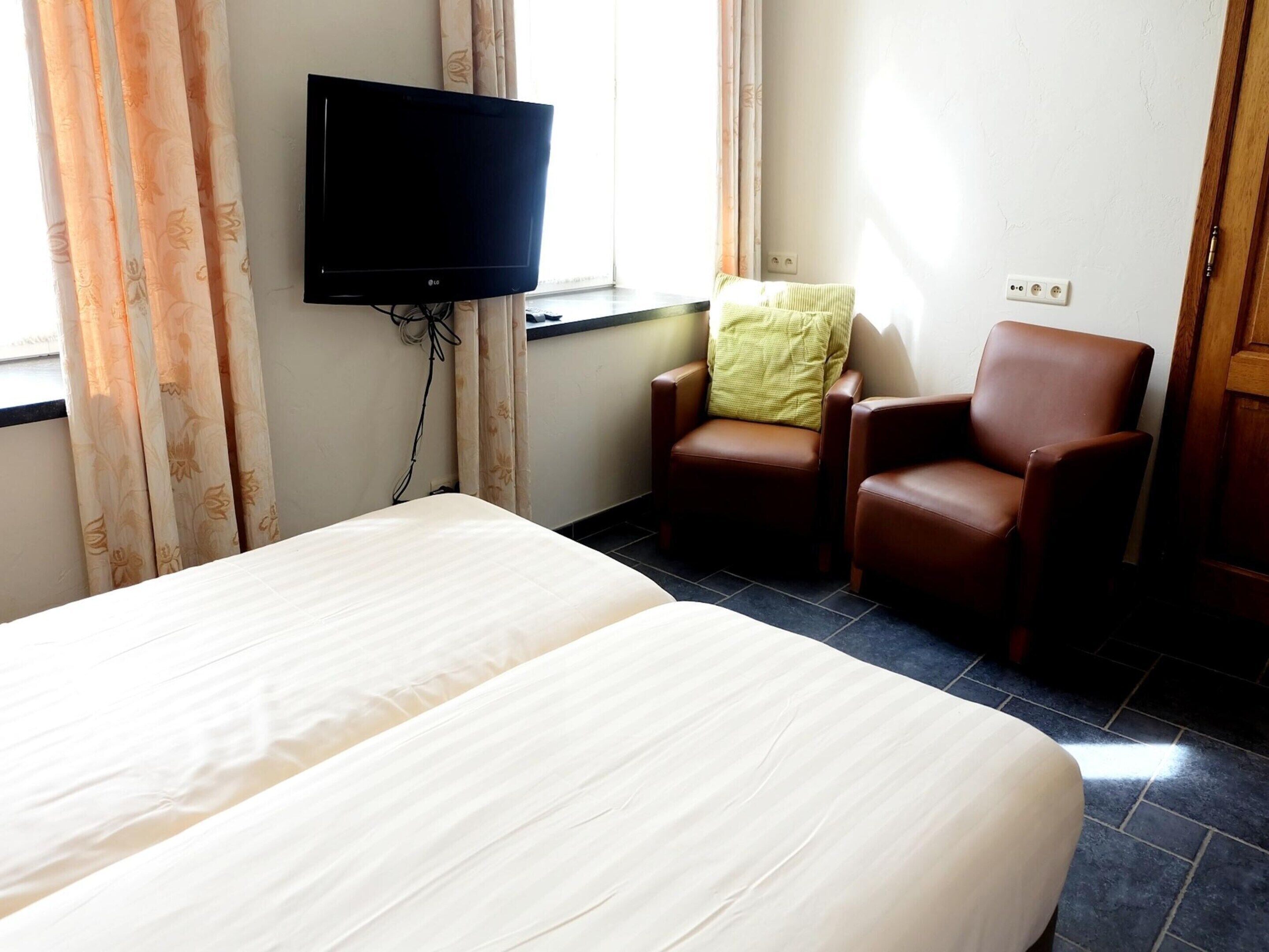 Quarto de hotel com cama, televisão na parede, uma cadeira e janela com cortinas.