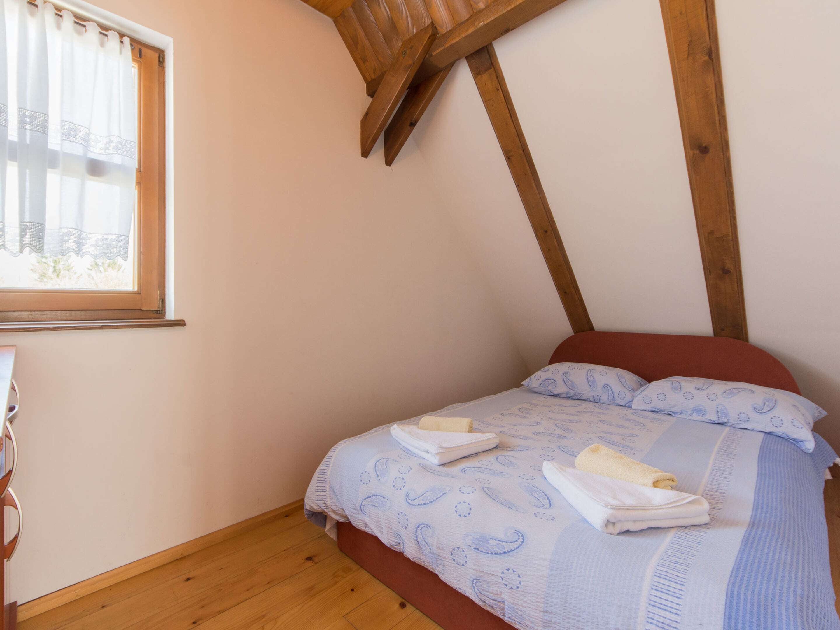 A bedroom with a wooden ceiling, a bed with a patterned duvet, and a window with sheer curtains.
