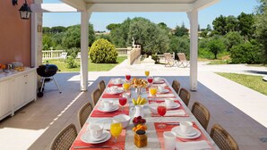 An outdoor dining area with a long table set for breakfast, surrounded by wicker chairs, and a barbecue grill on the left. - Belvilla by OYO Gira Sole (Syracuse)