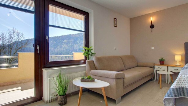Un salon moderne avec un canapé beige, une petite table ronde, une plante en pot et une vue sur une zone boisée par une grande fenêtre.