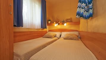 Quarto estilo cabana com duas camas de solteiro, cabeceiras de madeira, uma janela com cortinas leves, uma prateleira fixada na parede com objetos decorativos e cortina azul estampada.