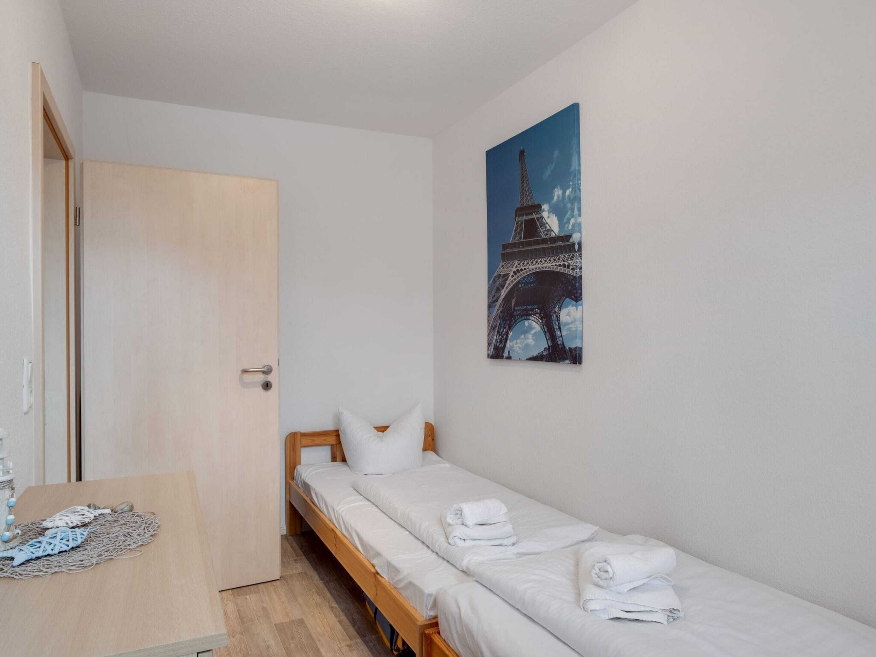 A small, single-bed room with a wooden bed, white bedding, a door, a small table with a decorative item, and a painting of the Eiffel Tower on the wall.