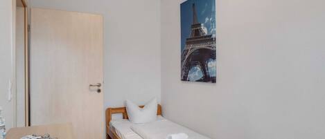 A small, single-bed room with a wooden bed, white bedding, a door, a small table with a decorative item, and a painting of the Eiffel Tower on the wall.