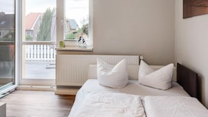 A bedroom with a bed, white pillows, a radiator, and a window with a view of houses and trees. - Snug Apartment in Kalkhorst with Terrace near City Center (Kalkhorst)