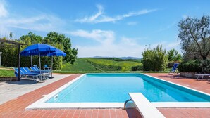 A rectangular swimming pool with a white edge, surrounded by red brick flooring. There are blue lounge chairs and umbrellas, and a white lounge chair is visible in the foreground. - Scenic Farmhouse in Peccioli with Swimming Pool near Lakes (Peccioli)