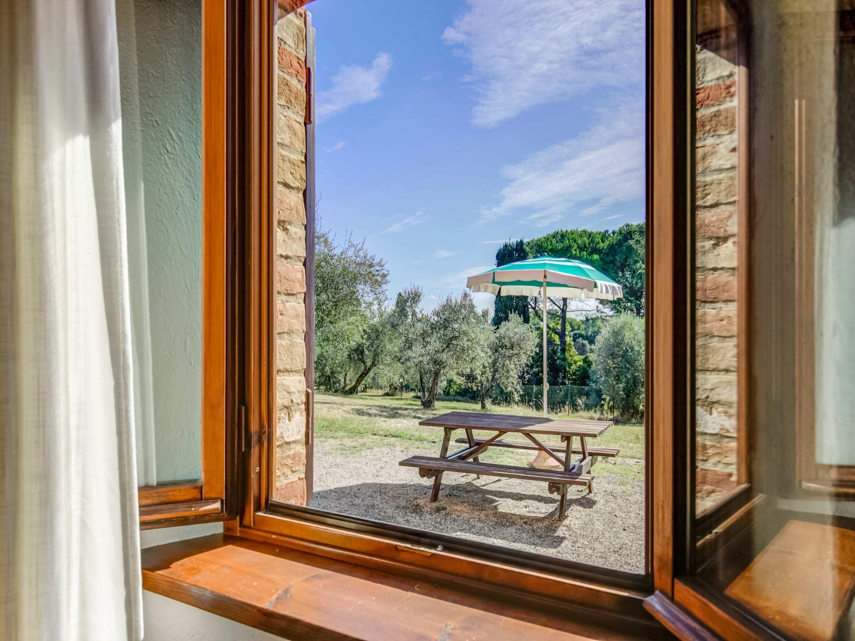 A view through a window with a wooden frame, a picnic table, and a green umbrella in an outdoor area with trees and a clear sky.