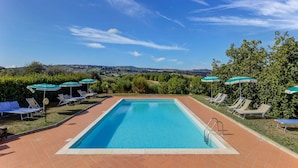 A rectangular swimming pool with a clear blue water surface, surrounded by a tiled deck. There are several lounge chairs and umbrellas around the pool, and a view of the countryside in the background. - Cozy Farmhouse With Pool (Gambassi Terme)