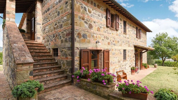 A stone building with a wooden staircase leading to a porch, surrounded by a well-maintained garden with flowering plants. - Belvilla by OYO Piandigoro - Le Querce (Radicofani)
