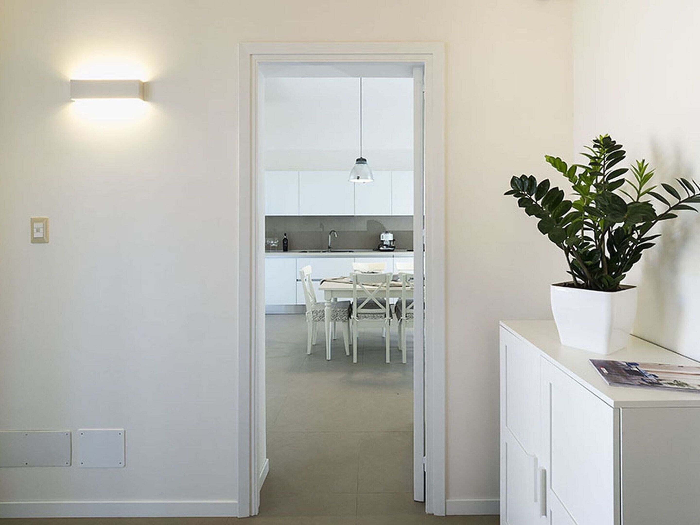Corredor que dá para uma cozinha com armários brancos, uma mesa de jantar com cadeiras e uma planta em vaso sobre um armário.