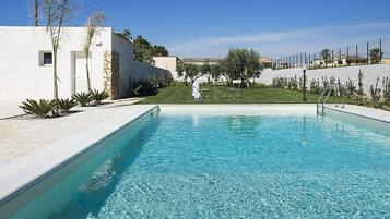 Uma piscina com água azul cristalina, rodeada por um edifício branco e uma área gramada com cerca.
