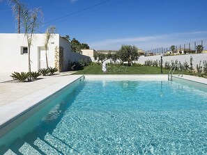 A swimming pool with clear blue water, surrounded by a white building and a grassy area with a fence.