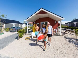A family with a child holding a beach ball in front of a red house with a porch. - Fully Furnished Tiny House near Beach (Maurik)