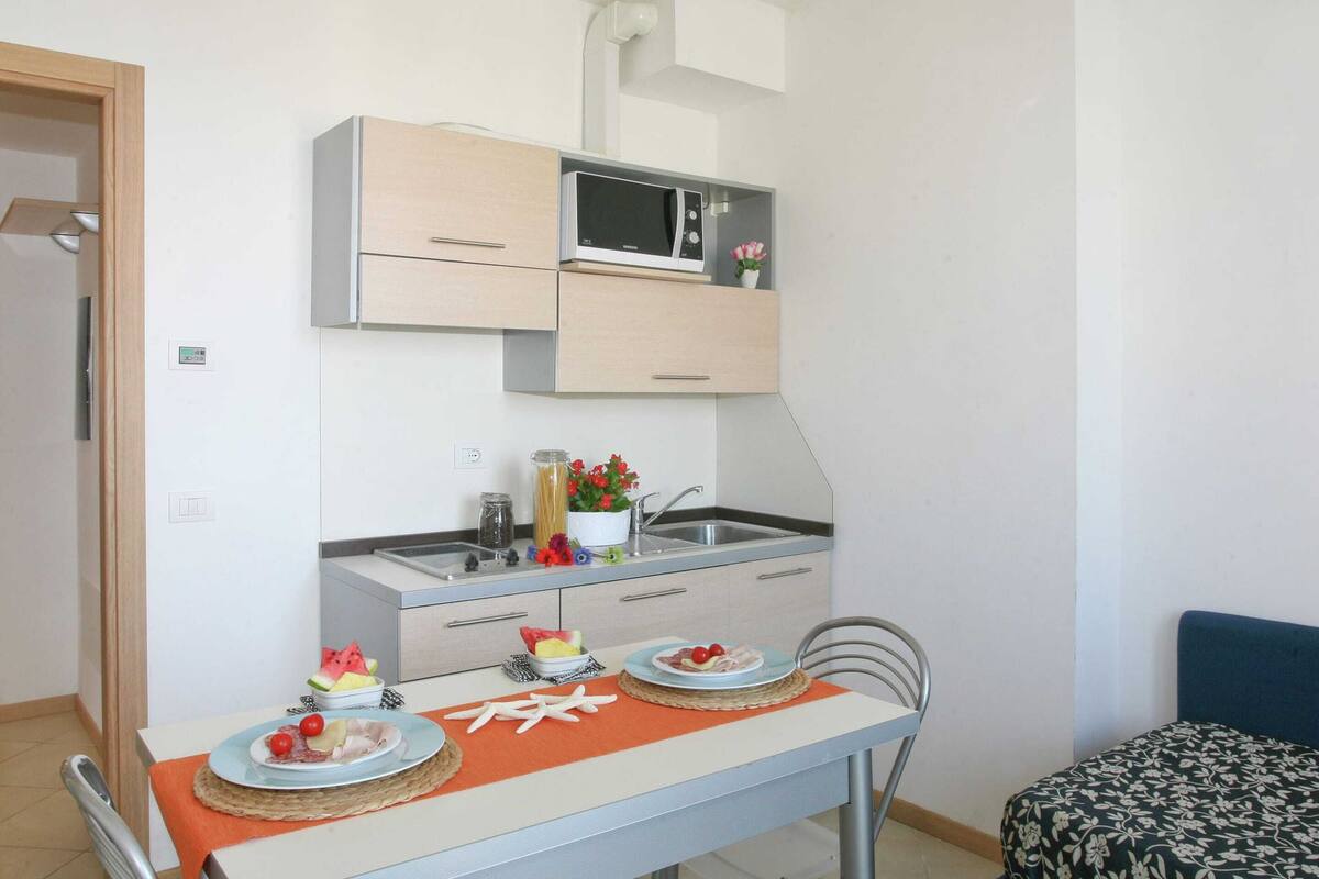 A compact kitchen with a microwave, sink, and cabinets. There is a dining table with plates and a chair.