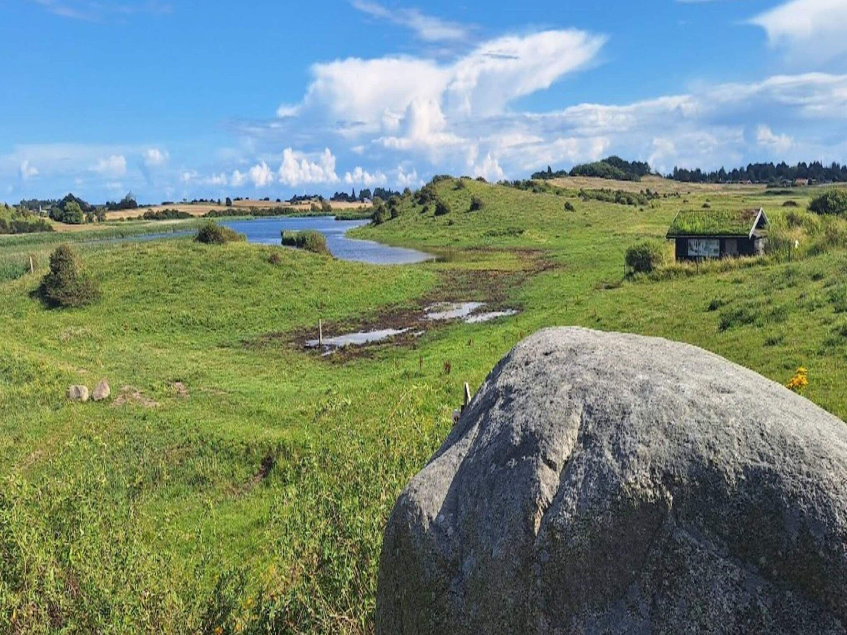 Un paysage naturel avec un grand rocher au premier plan, un petit plan d’eau et, au loin, une colline sur laquelle se trouve une construction.