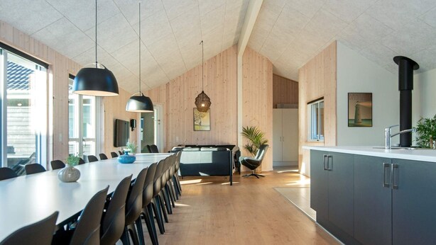 A modern kitchen with wooden walls, a dining area with a long table and chairs, and a kitchen island. - Holiday Home in Nørre Nebel (Norre Nebel)