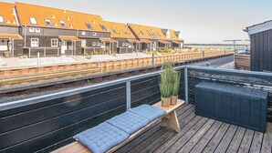 A wooden deck with a bench and potted plants, overlooking a row of buildings with orange roofs and a railway track. - Holiday Home in Juelsminde (Juelsminde)