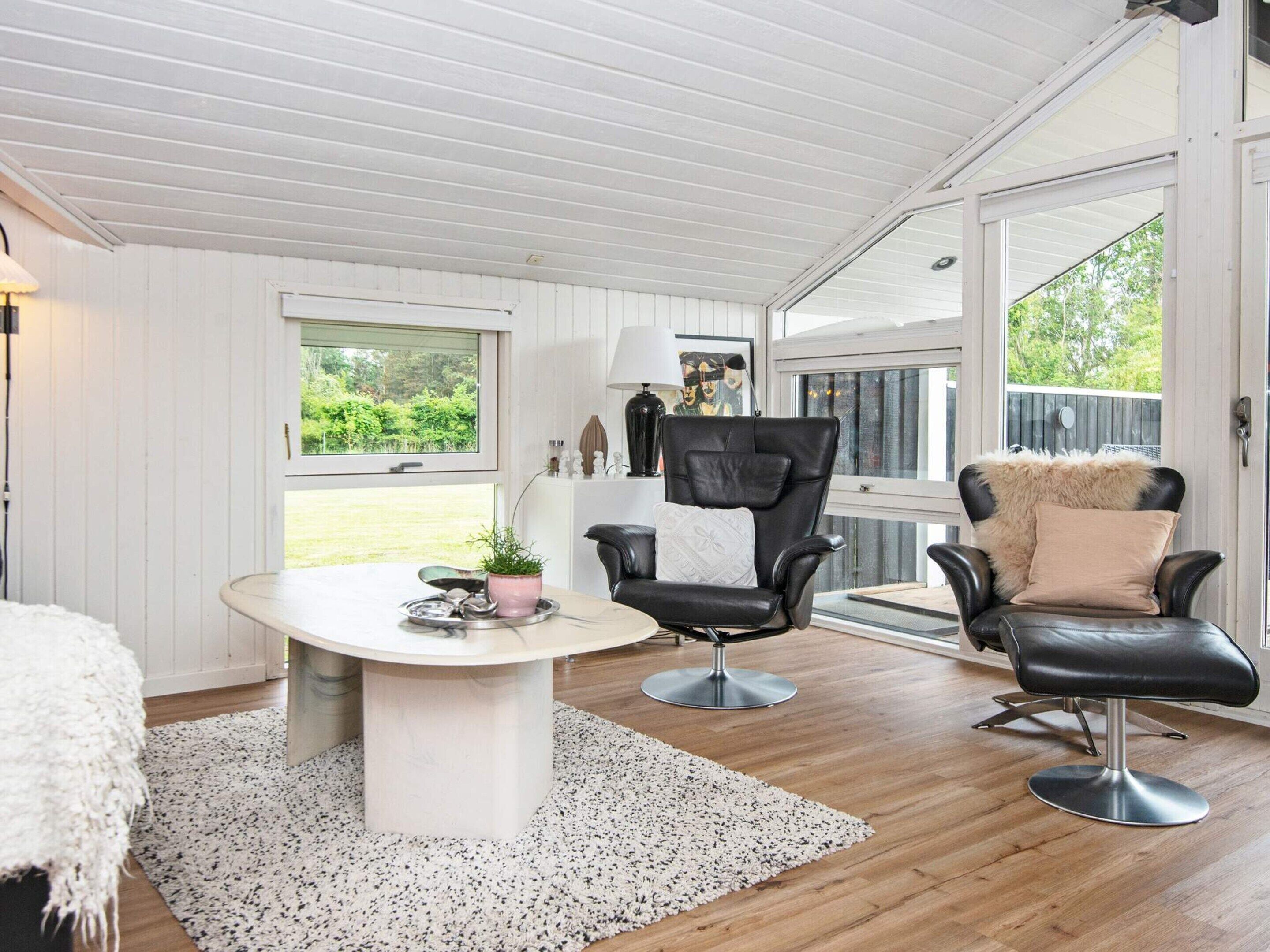 Een kamer met een wit plafond, een ronde tafel met een witte tafelkleed, twee zwarte lederen fauteuils met kussens, een wit vloerkleed en een raam met uitzicht op groen.