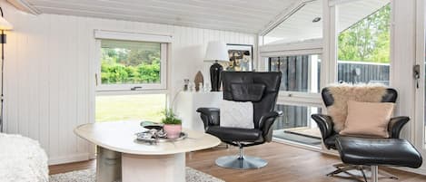 Een kamer met een wit plafond, een ronde tafel met een witte tafelkleed, twee zwarte lederen fauteuils met kussens, een wit vloerkleed en een raam met uitzicht op groen.