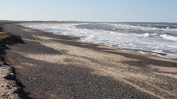 A beach with pebbles and sand, waves crashing, and a clear sky.
