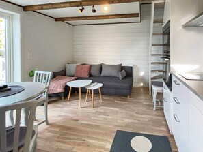 Una cocina moderna con armarios blancos, una zona de comedor con mesa y sillas blancas, un sofá gris con cojines, y piso de madera.