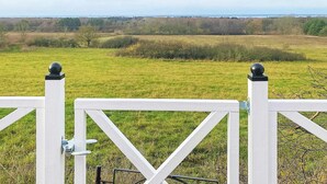 A white fence with a gate, a grassy field, and a distant view of trees and a body of water. - 4 Person Holiday Home in Farjestaden (Farjestaden)