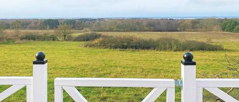 A white fence with a gate, a grassy field, and a distant view of trees and a body of water.