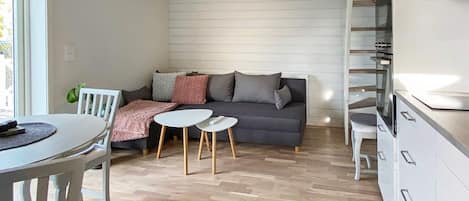Una cocina moderna con armarios blancos, una zona de comedor con mesa y sillas blancas, un sofá gris con cojines, y piso de madera.