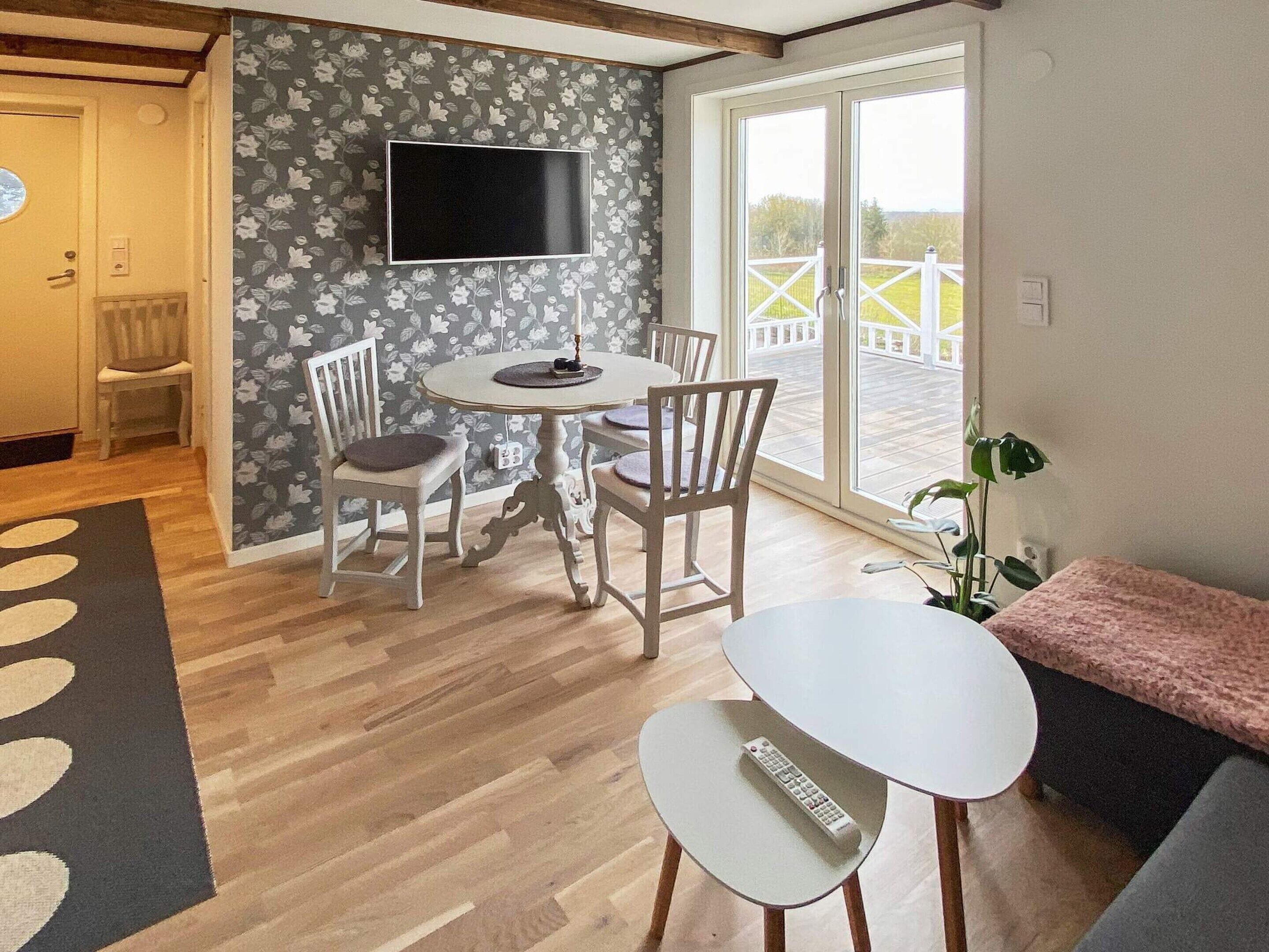 A dining area with a table and chairs, a TV mounted on the wall, and a large window with a view.