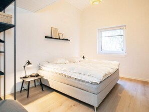 A minimalist bedroom with a bed, a small table, a lamp, a shelf with items, and a window. - Holiday Home in Sæby (Sæby)