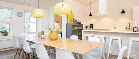 A modern kitchen with a wooden table, white chairs, hanging wicker lamps, and a visible clock.