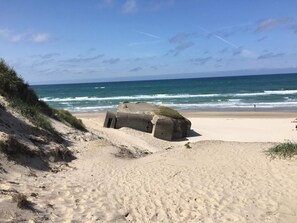 En sandstrand med en betongkonstruktion, eventuellt en bunker, och havsvågor i bakgrunden.