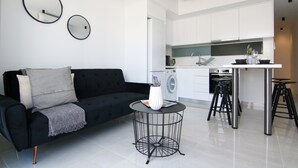 Uma sala de estar moderna com um sofá preto, uma mesa de centro redonda e uma cozinha com armários brancos e uma pia de aço inoxidável.