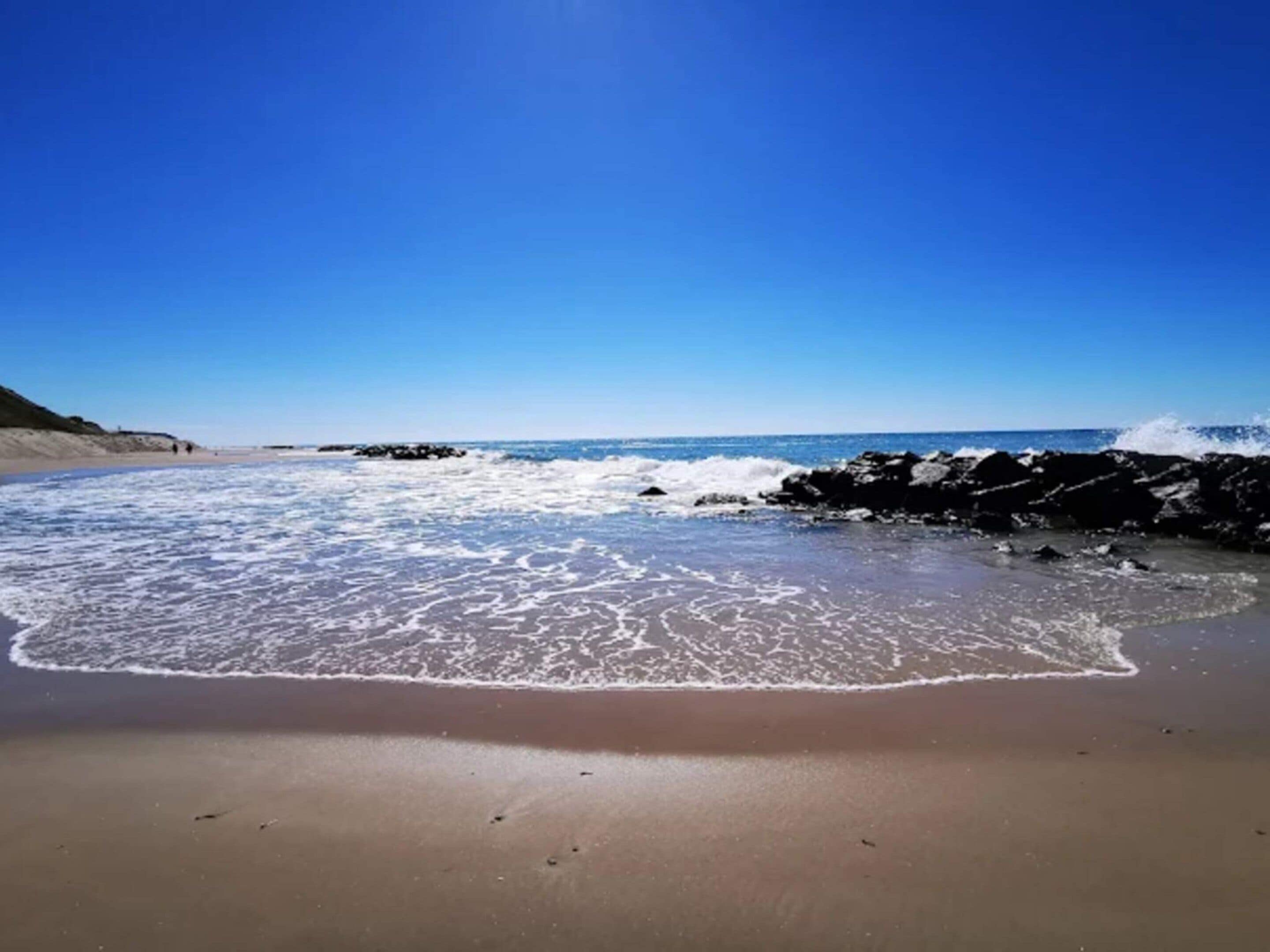 A beach with waves crashing onto the shore, a clear sky, and a rocky outcrop in the distance.