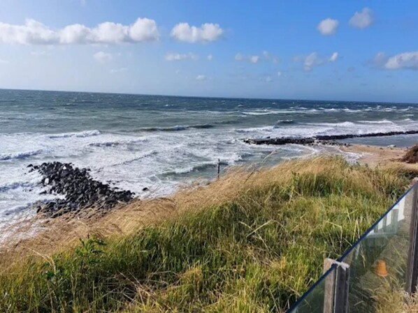 A coastal landscape with waves crashing onto a rocky shore, a grassy area, and a clear sky.
