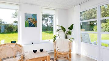 A bright room with large windows, a wooden table, two wicker chairs, and a colorful rug.