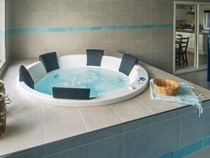 A hot tub with a blue water feature, surrounded by chairs, in a room with tiled walls and a visible kitchen area. - Holiday Home in Væggerløse (Vaeggerlose)