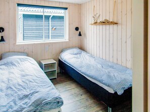 A small room with two single beds, a window with blinds, a bedside table, and a shelf with decorative items. - Holiday Home in Væggerløse (Vaeggerlose)