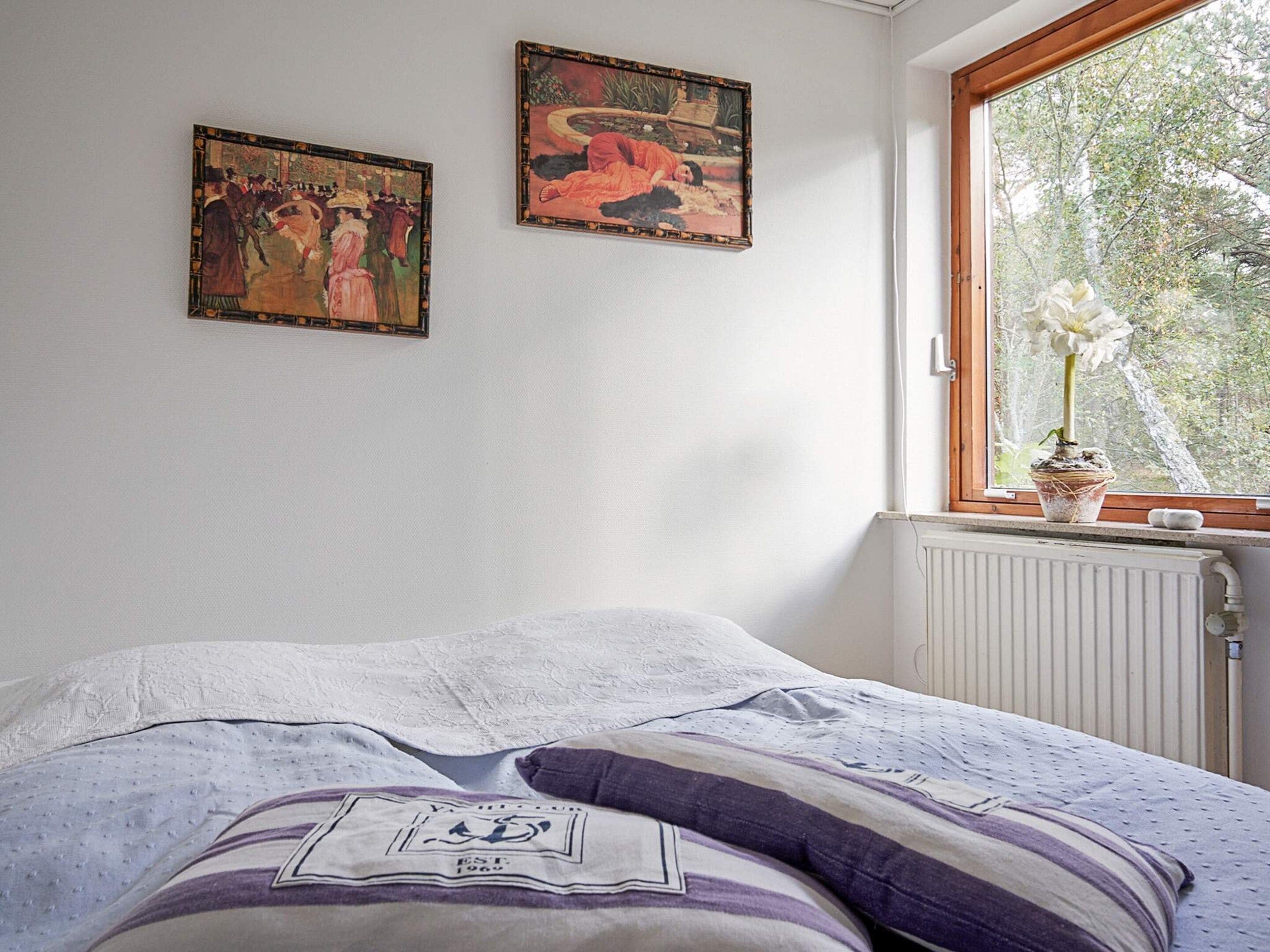 A bedroom with a bed, two paintings on the wall, and a window with a view of trees.