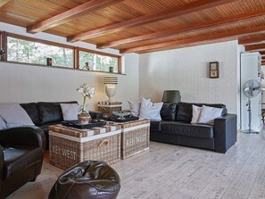 Uma sala de estar moderna com teto de madeira, parede de tijolos brancos e uma janela grande. Há dois sofás de couro preto, uma mesa de centro de vime e um ventilador de pé.