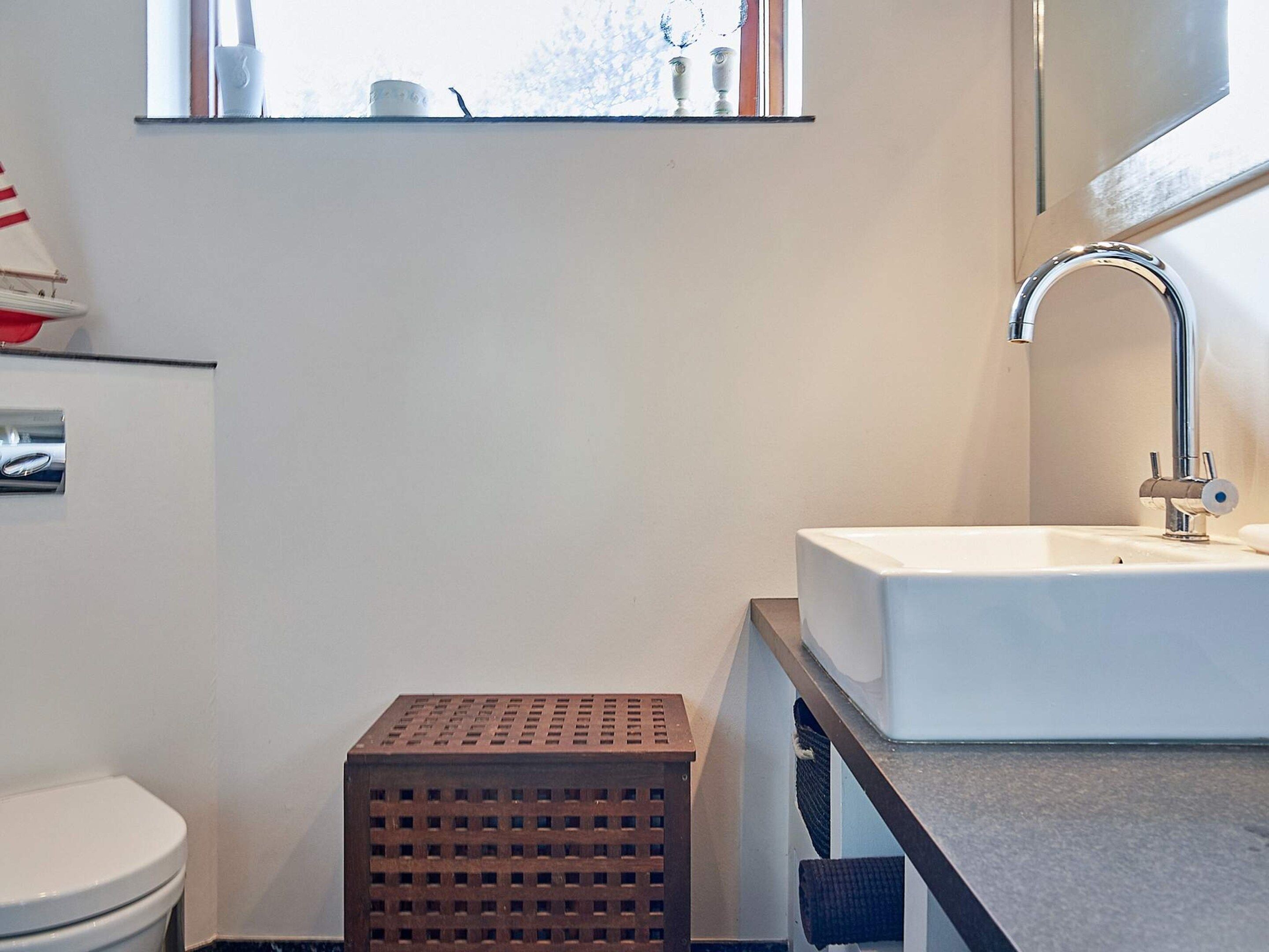 A bathroom with a white sink, a modern faucet, a small wooden stool, and a window with a view of trees.