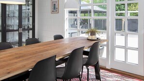 Una cocina moderna con una mesa de madera, sillas negras, luces colgantes y vistas al patio trasero a través de amplios ventanales.