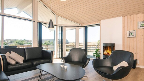 A modern living room with a black leather sofa, a black armchair, and a coffee table. There is a fireplace with a visible fire, large windows, and a wooden ceiling.