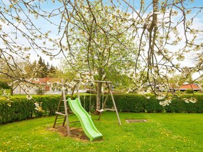 A playground with a green slide and swings, surrounded by a grassy area and flowering trees.