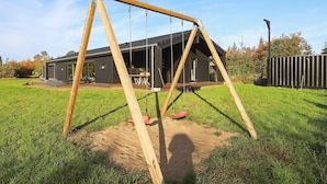 A wooden swing set in a grassy yard with a modern house in the background. - Holiday Home in Sæby (Sæby)