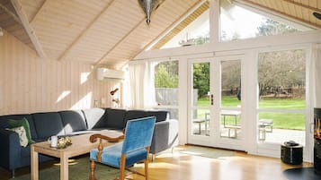 Una habitación con techo de madera, una silla azul, un sofá, una mesa y un amplio ventanal con vista a un jardín.