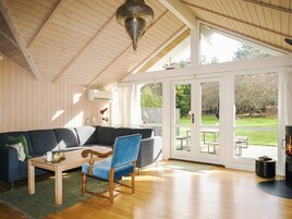 Una habitación con techo de madera, una silla azul, un sofá, una mesa y un amplio ventanal con vista a un jardín.