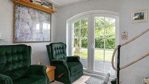 A room with two green upholstered chairs, a wooden table, a large framed painting, and a view through a glass door to a deck. - 6 Person Holiday Home in Lokken (Lokken)